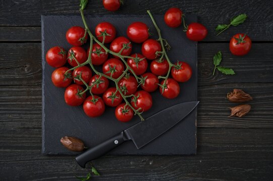 Fresh red cherry tomatoes arranged on a dark wooden surface with a rustic feel, accompanied by a black kitchen knife on a slate serving tray. - Powered by Adobe