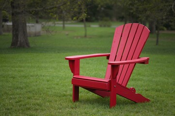 Crimson resin Muskoka Adirondack seat set on lush lawn in a park on a warm spring or summer day, representing leisurely outdoor getaway.
