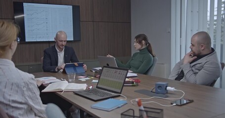 Business team analyzing financial data in a meeting, using laptops, tablets, and a large screen display. - Powered by Adobe