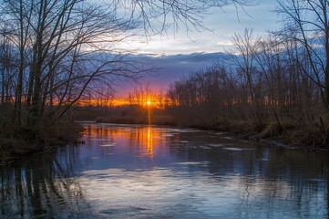 A tranquil river meanders quietly as daylight fades, framed by leafless trees and a vibrant sky, evoking calm and introspection.