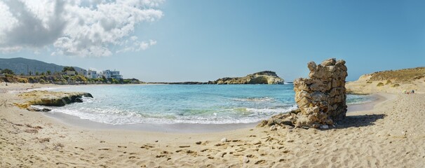 A sunny beach with clear blue water and a sandy shore. Clouds and houses in the background create a calm atmosphere, Arkasa, Karpathos Island, Greece