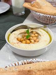 A bowl of hummus topped with chickpeas and olive oil, garnished with a sprig of mint. Served with pita bread and a basket of breadsticks.