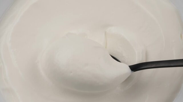 Close-up of a black spoon dipping into thick white yogurt or cream. Bowl with yogurt or kefir, lactic acid product dairy concept. Smooth texture, fresh lactose free in morning. Breakfast background