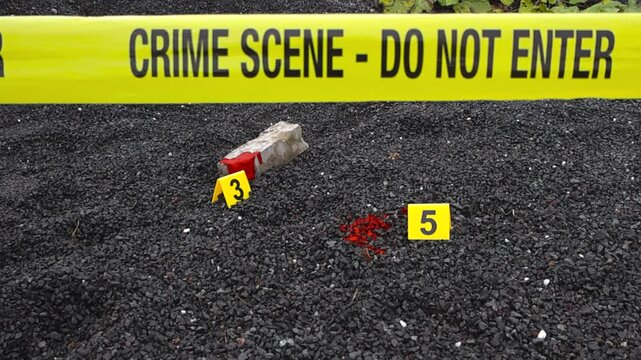 Yellow police line crime scene forensic investigation tape ribbon in front of a yellow crime markers that are placed next to blood stain and a bloody brick or blunt force object on dark gravel. Bokeh.