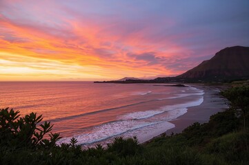 Obraz premium At dawn, the tranquil sea mirrors the vivid hues of the sky, casting light on the scenic shore where ocean waves meet the hills.