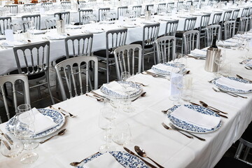Formal dinning setup of chairs, long tables with tablecloths, plates, cutlery and glasses. Wide angle, interior setting, top down, no people