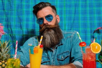 Man with a long beard a brutal humorous hipster in denim surrounded by tropical cocktails on a colorful background