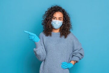 Mixed race young woman with brown hair wearing a mask and gloves gestures to the side while showcasing a product against a blue background