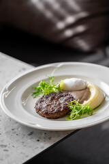 Gourmet dish with grilled meat patty, mashed potatoes, creamy topping, brown sauce, and fresh herbs on a white plate. Minimalist presentation, fine dining style.
