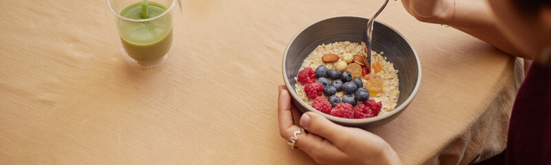 Website header shot of young adult woman eating oatmeal topped with blueberries, raspberries, almonds and orange slices while sitting at table with green juice nearby, holding bowl with both hands