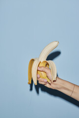Caucasian young adult woman hand holding partially peeled banana against blue background, fingers...