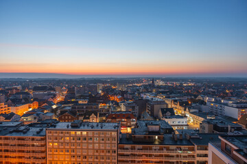 The town lights glow as night falls, with the sunset fading behind the horizon. Streets and buildings illuminate under the twilight sky.