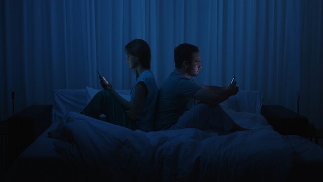 A couple sits back to back in bed at night using smartphones