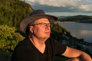 Close-up portrait of a thoughtful man in hat and glasses enjoying sunset light near the lake. Warm...