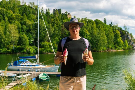 Man in casual summer outfit stands near a peaceful lakeside marina, holding two ice cream cones under bright sunlight. Yachts and green forest create a relaxed vacation vibe, capturing leisure, nature - Powered by Adobe
