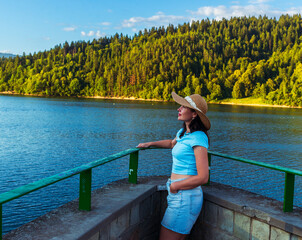 Young woman in blue top and straw hat leans on a stone railing, gazing over calm lake water surrounded by green forest. A serene summer scene evoking freedom, relaxation, and mindfulness in nature.