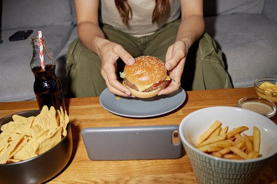 Young adult Caucasian woman sitting at table holding hamburger with both hands, watching smartphone screen, surrounded by bowl of fries, tortilla chips, dipping sauces, and bottle of soda