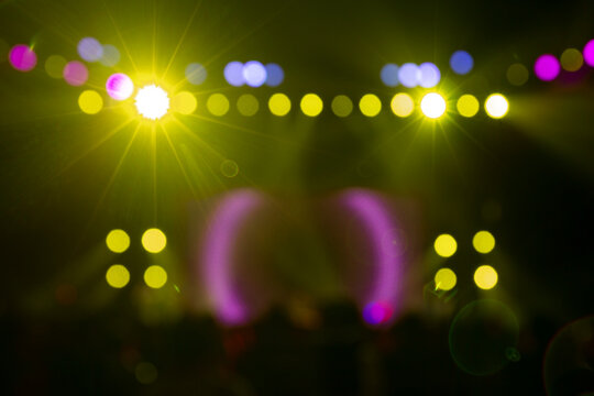 Abstract blurred background with vibrant stage lights, showcasing glowing yellow spots, purple arcs, and blue bokeh effects. Dynamic lens flares add energy to this concert or event backdrop.