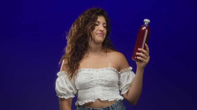 Young hispanic woman holds a bottle of juice and shows bare shoulder in a blue studio; curiosity detox refreshment.