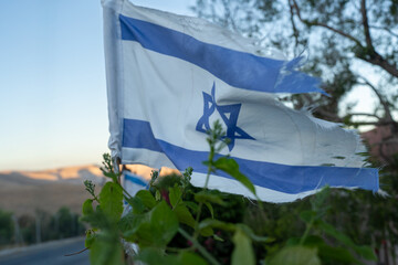 A frayed Israeli flag flutters gently in the breeze, surrounded by greenery. The sun sets behind distant hills, casting a warm glow on the landscape and flag, symbolizing resilience.