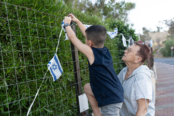 Grandmother assists a boy as he climbs a fence to hang small Israeli flags. Senior woman helps kid hang an Israeli flag on garden fence, showing love, unity, pride before Israel Independence Day.