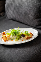 Gourmet dish with leafy greens and pastry served on a white plate, placed on a dark fabric surface. Elegant food styling in an unconventional, casual setting.