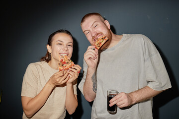 Caucasian young woman and Caucasian young man eating pizza together, smiling and looking at camera, man holding glass of dark beverage, casual setting, camera flash