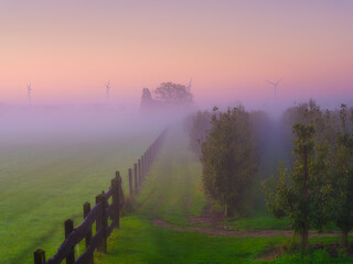 Garden during foggy sunrise. Wind turbine in the fog. Renewable and natural energy sources. Field and meadow. Fence on the field. Photo for background, wallpaper, postcards.