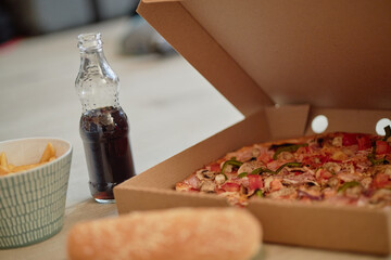 Open pizza box displaying freshly baked pizza with visible toppings next to glass bottle of soda, bowl of fries and sandwich bun on light table surface, food arranged for meal