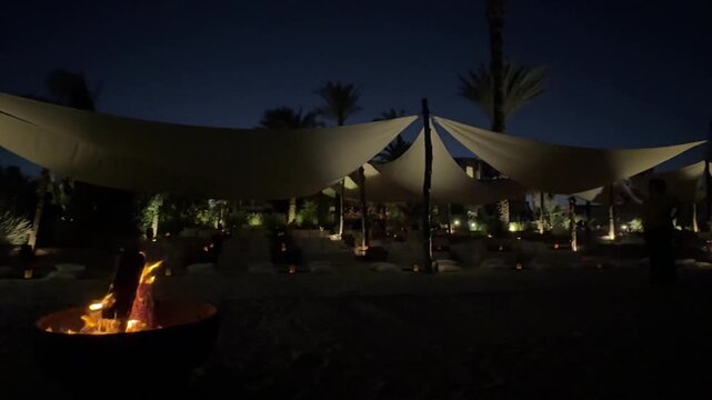 A night scene with a campfire, people sitting around, enjoying warmth and conversation. Tents and outdoor items visible, creating a cozy atmosphere. Dark sky, trees, and nature elements in the backgro