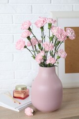 Beautiful carnation flowers in vase and other decor on wooden cabinet near white brick wall indoors