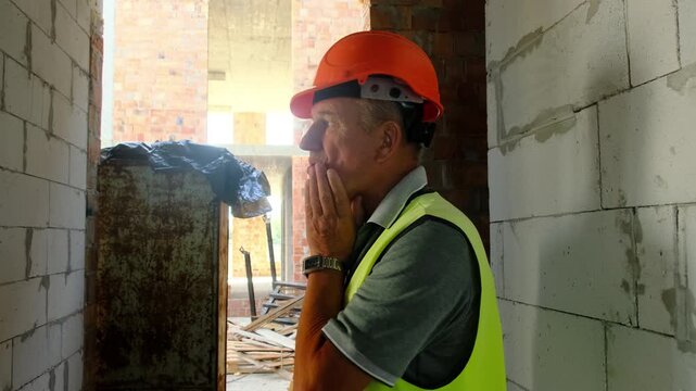 Construction worker in safety vest thinks about work process. Experienced constructor touches face with hand and inspects building analyzing results