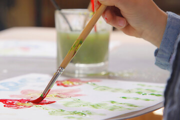A child’s hand painting with a brush, creating colorful artwork on paper. A glass of green water with brushes in the background