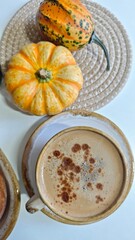 Cup of frothy cappuccino with cinnamon on a ceramic saucer beside decorative pumpkins on a white background
