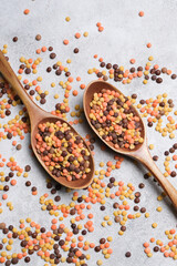 Colorful lentils in two wooden spoons scattered on a grey textured surface, top view