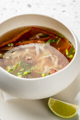 Asian noodle soup with beef, rice noodles, chili, green onion, and lime wedge served in a white bowl on a plate. Minimalist presentation on light speckled background.
