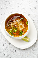 Asian noodle soup with beef, rice noodles, chili, green onion, and lime wedge served in a white bowl on a plate. Minimalist presentation on light speckled background.