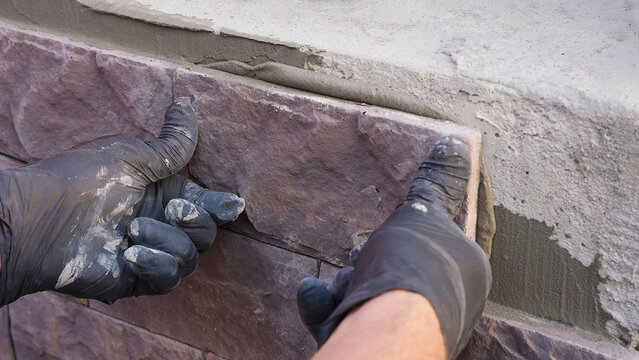 Close-up of masonry work as a construction worker installs decorative stone on a wall with mortar. Hands in gloves placing materials during facade renovation and repair