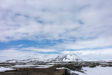 snow covered Mt. Hl&iacute;&eth;arfjall emerging from sorrounding lava field under cloudy blue sky Lake M&yacute;vatn, North Iceland