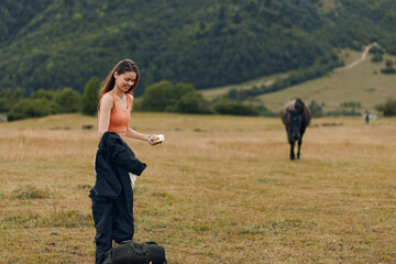 A young woman stands in a wide grassy field with a horse in the distance, adjusting her gear and...