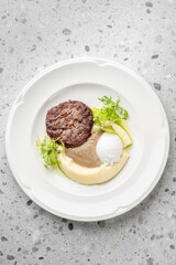 Gourmet dish with meat patty, poached egg, mashed potatoes, brown sauce, avocado slices, and frisée lettuce on white plate. Elegant plating, fine dining presentation.