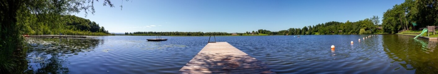 Fototapeta premium Heratinger See, Ibm, Peaceful Lake with Wooden Dock and Recreational Features Surrounded by Trees – Sunny Day in Upper Austria 