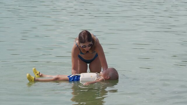 A caring woman in a blue bikini helps a young child, wearing a floatation vest and yellow water shoes, happily float on their back in calm, light blue water, creating a tender moment of learning