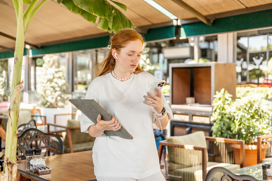 Young European woman with red hair holding laptop and smartphone in outdoor cafe, working remotely and studying online.