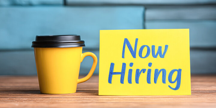 A bright yellow sign reads "Now Hiring" next to a yellow coffee cup, suggesting a job opportunity in a cozy environment.