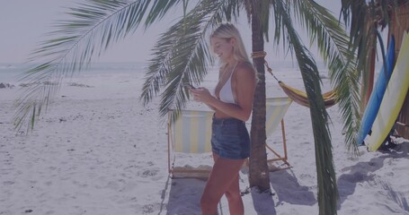 Holding smartphone woman wearing bikini top and denim shorts at beach with surfboards, copy space