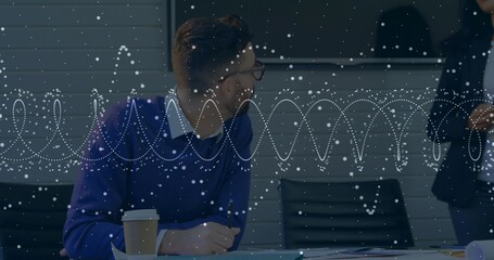 Writing man in blue sweater glancing at blazer-clad woman in meeting room, with smartphone