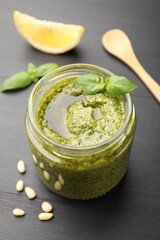 Fresh pesto sauce in jar, spoon and ingredients on black wooden table, closeup