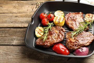 Pan with grilled beef steaks, rosemary and vegetables on wooden table, closeup. Space for text