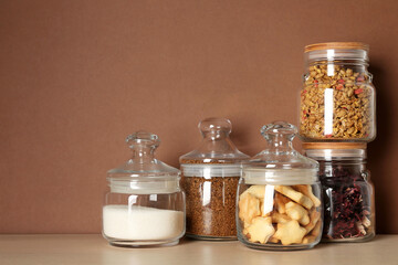 Glass jars with different products on wooden table against brown background. Space for text